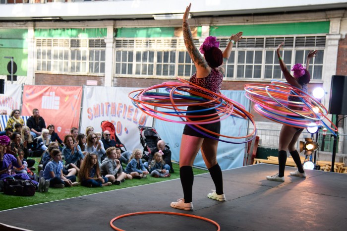 Image of two hula hoopers on stage to an audience of kids in the background. They both have their arms up in the air in a joyous pose with loads and loads of hoops around their waists.